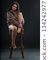 brunette woman sits with one leg curled under her on a chair. She wears a hoodie and looks directly at the viewer in a dark room with soft lighting. Her expression is relaxed. 134242977