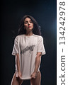 brunette woman stands in a studio with soft light. She holds her white T shirt with both hands looking directly at the camera. The setting is dim and focused on her. 134242978
