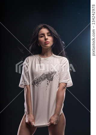 brunette woman stands in a studio with soft light. She holds her white T shirt with both hands looking directly at the camera. The setting is dim and focused on her. 134242978