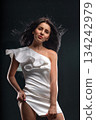 A brunette woman stands confidently wearing a short white dress. Her hair flows freely as she poses against a dark backdrop highlighting her fashion choice and personality. 134242979