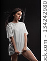 brunette stands in a white T shirt against a dark background. She looks thoughtful while holding the hem of her shirt. The studio light highlights her features and creates a dramatic atmosphere. 134242980