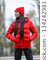 brunette woman stands at the stadium with a red jacket and matching hat. She has her hands in her pockets and looks confidently ahead. The atmosphere is cloudy and cool. 134242981