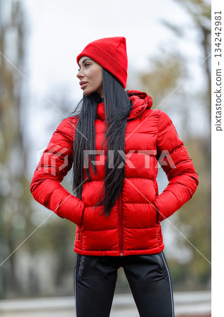 brunette woman stands at the stadium with a red jacket and matching hat. She has her hands in her pockets and looks confidently ahead. The atmosphere is cloudy and cool. brunette woman stands at the stadium with a red jacket and matching hat. She has her hands in her pockets and looks confidently ahead. The atmosphere is cloudy and cool. 134242981