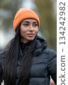 brunette woman stands at a stadium wearing a black jacket and an orange hat. She looks around while attending an outdoor event with many people and activities happening. 134242982