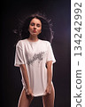 brunette stands with her hands holding the bottom of a white T shirt. She is in a studio with dark background and bright lights showcasing a confident pose. The time appears to be night. 134242992