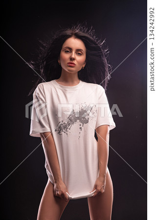 brunette stands with her hands holding the bottom of a white T shirt. She is in a studio with dark background and bright lights showcasing a confident pose. The time appears to be night. 134242992