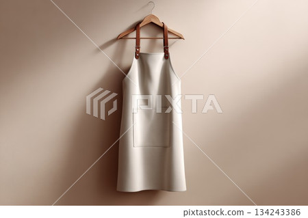 White apron mockup, simple uniform hanging on a wooden hanger with leather straps against a smooth beige wall. Blank apparel template, clean design for a commercial kitchen or coffee shop. 134243386