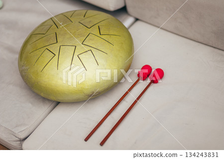 Golden handpan drum with geometric sound holes rests on a sofa, by two vibrant red mallets Golden handpan drum with geometric sound holes rests on a sofa, by two vibrant red mallets 134243831