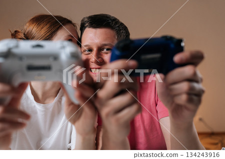 Cheerful young couple enjoying home entertainment, holding wireless video game controllers, bonding over competitive or cooperative gameplay, having fun together, leisure activity at home. 134243916