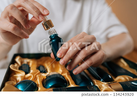 Woman hands carefully holding dropper with liquid serum above a small bottle, part of a luxurious cosmetic gift set nestled in golden fabric, emphasizing beauty rituals and personal care. 134243920