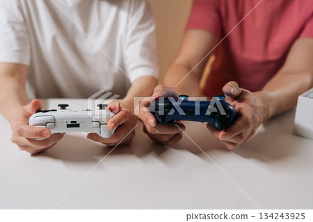 Two people holding white and blue wireless controllers, engaging in gaming session sitting at white table, enjoying modern entertainment and companionship. Two people holding white and blue wireless controllers, engaging in gaming session sitting at white table, enjoying modern entertainment and companionship. 134243925