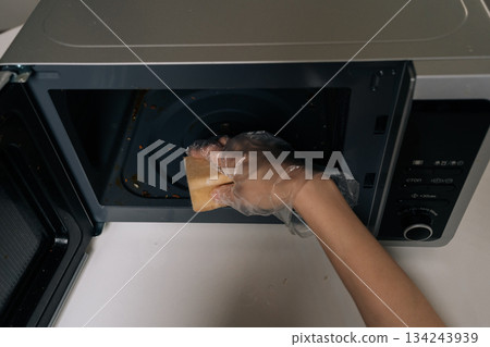 Close-up of female hands in clear disposable gloves scrubbing inside of microwave with yellow sponge, close-up of routine kitchen cleaning and appliance hygiene maintenance. 134243939