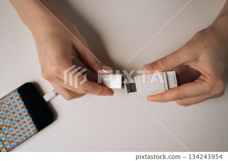 Top view of young adult woman hands connecting USB flash drive to smartphone using adapter cable sitting at white table, transferring data or charging device, close-up. Concept of modern technology. Top view of young adult woman hands connecting USB flash drive to smartphone using adapter cable sitting at white table, transferring data or charging device, close-up. Concept of modern technology. 134243954