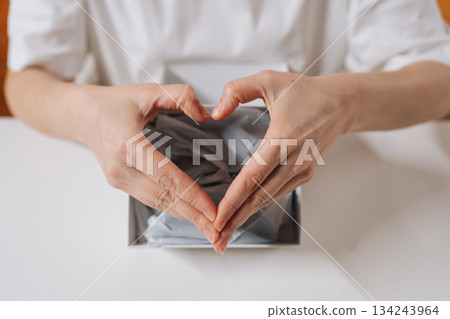 Woman hands forming prominent heart shape position over open white gift box, symbolizing love, care and gratitude during unpacking process of special present, close-up. 134243964