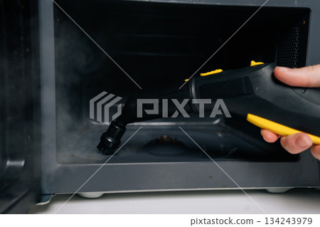 Close-up of person cleaning microwave oven interior with steam cleaner appliance for hygiene and disinfection, removing grease and food residue from appliance surface. Concept of kitchen hygiene. Close-up of person cleaning microwave oven interior with steam cleaner appliance for hygiene and disinfection, removing grease and food residue from appliance surface. Concept of kitchen hygiene. 134243979