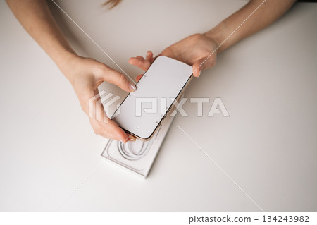 Top view of female hands unpacking new mobile phone from box on white table, eagerly holding device and preparing for exciting first-time setup experience. Concept of smartphone unboxing process Top view of female hands unpacking new mobile phone from box on white table, eagerly holding device and preparing for exciting first-time setup experience. Concept of smartphone unboxing process 134243982