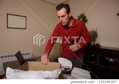 Man Packing Box At Home With Clothes In Living Room, Preparing Shipment Man Packing Box At Home With Clothes In Living Room, Preparing Shipment 134244167