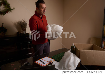 Man Unboxes Package At Home With Christmas Decor, Box, And Plastic Wrap 134244171
