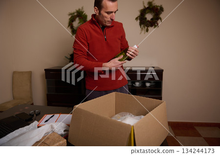 Man in Red Sweatshirt Unpacking Bottle from Box in Cozy Home Kitchen Setting Man in Red Sweatshirt Unpacking Bottle from Box in Cozy Home Kitchen Setting 134244173