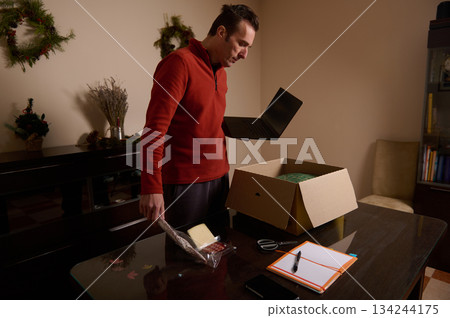 Man Unboxing Christmas Gift At Home With Laptop And Box On Table Man Unboxing Christmas Gift At Home With Laptop And Box On Table 134244175