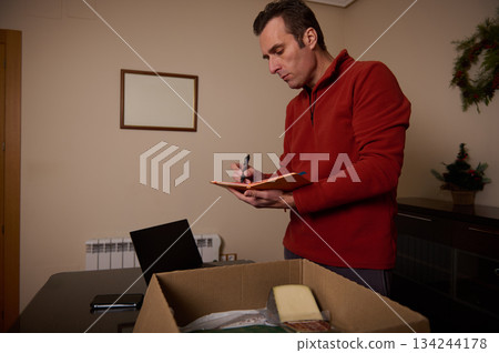 Professional Man in Red Sweater Notes Inventory While Packing Box at Home Office 134244178