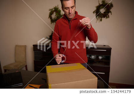 Man In Red Sweater Seals Cardboard Box With Tape In Cozy Home Office 134244179