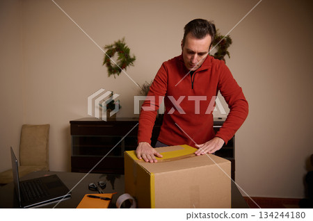Man In Red Sweater Unpacks Package At Home Desk Amid Holiday Decor 134244180