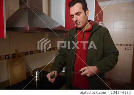 Man Cooking In Modern Kitchen With Knife, Cutting Board, And Focused Expression Man Cooking In Modern Kitchen With Knife, Cutting Board, And Focused Expression 134244183