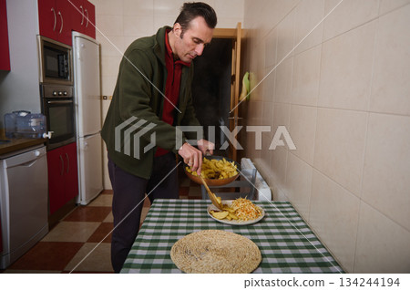 Man Serves Home-Cooked Dinner in Kitchen, Plating Potatoes and Fries on Checkered Table Man Serves Home-Cooked Dinner in Kitchen, Plating Potatoes and Fries on Checkered Table 134244194