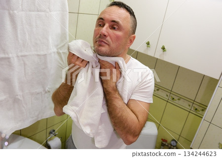 A blonde man with a beard drying his face with a white towel indoors, symbolizing hygiene and grooming in a bright, home setting. 134244623