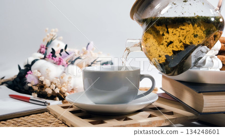 Fresh loose leaf tea flows from glass teapot into white cup, lemon slices and cookies with flowers add cozy and relaxing atmosphere  134248601