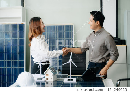 Two Asian engineers collaborate on a solar and wind energy project, discussing innovation and sustainability in a modern office setting. 134249623