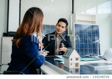 Two Asian engineers collaborate on a solar and wind energy project, discussing innovation and sustainability in a modern office setting. Two Asian engineers collaborate on a solar and wind energy project, discussing innovation and sustainability in a modern office setting. 134249720