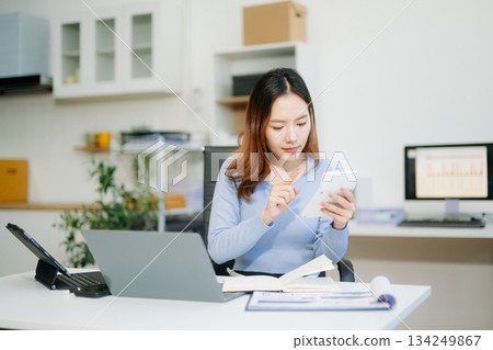 Asian woman smiling thinking planning writing in notebook, tablet and laptop working from home Asian woman smiling thinking planning writing in notebook, tablet and laptop working from home 134249867