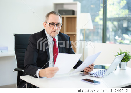 Confident businessman in suit, working on laptop and smartphone at office desk. Professional executive, leadership, strategy 134249942