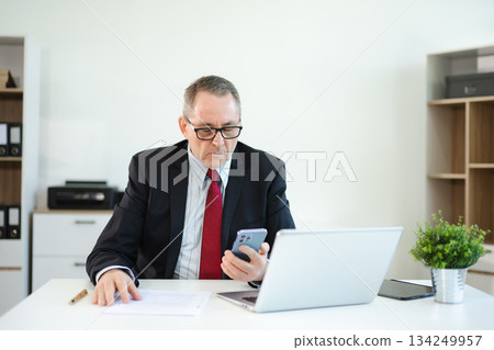 Confident businessman in suit, working on laptop and smartphone at office desk. Professional executive, leadership, strategy Confident businessman in suit, working on laptop and smartphone at office desk. Professional executive, leadership, strategy 134249957