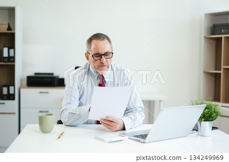 Senior businessman in formal attire using tablet and drinking coffee at modern office desk, working on finance, planning, or digital consulting tasks. 134249969
