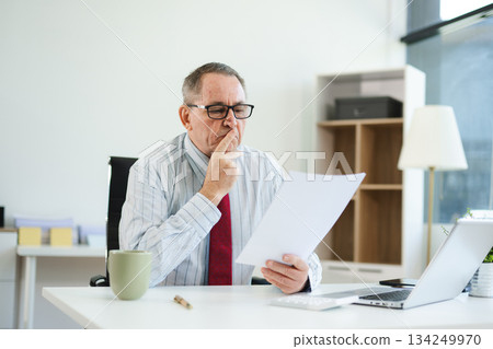 Focused senior executive analyzing printed documents at modern office desk with laptop and coffee, working on business strategy, finance, 134249970