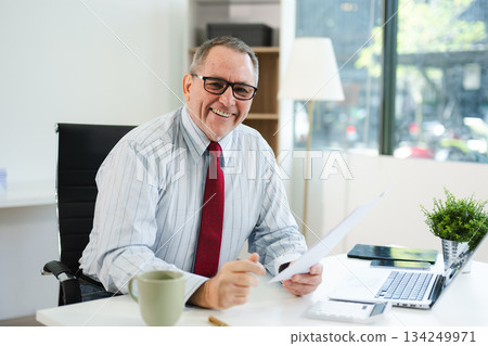 Focused senior businessman holding a document and using calculator at modern office desk, analyzing finance, budget, and corporate planning data with coffee beside. Focused senior businessman holding a document and using calculator at modern office desk, analyzing finance, budget, and corporate planning data with coffee beside. 134249971