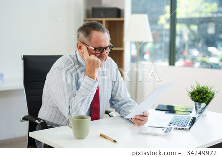 Focused senior executive analyzing printed documents at modern office desk with laptop and coffee, working on business strategy, finance, 134249972