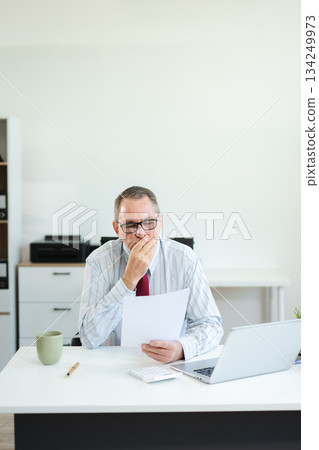 Focused senior executive analyzing printed documents at modern office desk with laptop and coffee, working on business strategy, finance, 134249973