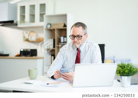 Focused senior businessman holding a document and using calculator at modern office desk, analyzing finance, budget, and corporate planning data with coffee beside. 134250033