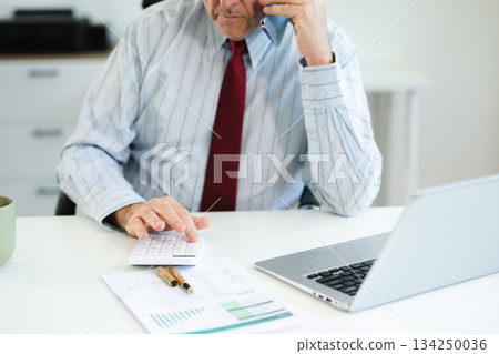 Senior businessman talking on smartphone while reviewing finance documents at modern office desk with laptop, multitasking on planning, communication, and business strategy. Senior businessman talking on smartphone while reviewing finance documents at modern office desk with laptop, multitasking on planning, communication, and business strategy. 134250036