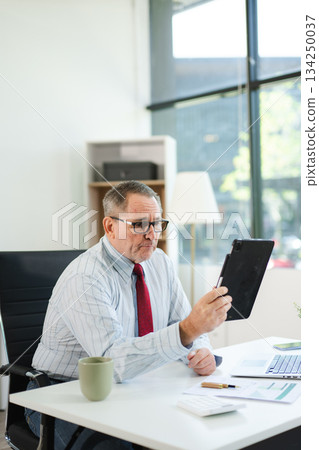 Focused senior businessman holding a document and using calculator at modern office desk, analyzing finance, budget, and corporate planning data with coffee beside. Focused senior businessman holding a document and using calculator at modern office desk, analyzing finance, budget, and corporate planning data with coffee beside. 134250037