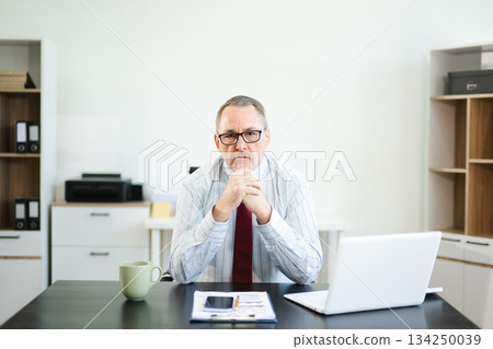 Frustrated senior businessman with smartphone gestures in confusion at office desk, reacting to bad news, tech issue, or business problem with laptop and reports. Frustrated senior businessman with smartphone gestures in confusion at office desk, reacting to bad news, tech issue, or business problem with laptop and reports. 134250039