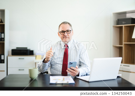 Smiling senior businessman in striped shirt and red tie gives a thumbs up at office desk, expressing confidence, approval, trust, and professional success. 134250043