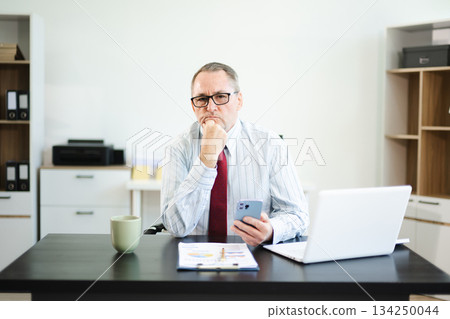 Frustrated senior businessman with smartphone gestures in confusion at office desk, reacting to bad news, tech issue, or business problem with laptop and reports. Frustrated senior businessman with smartphone gestures in confusion at office desk, reacting to bad news, tech issue, or business problem with laptop and reports. 134250044