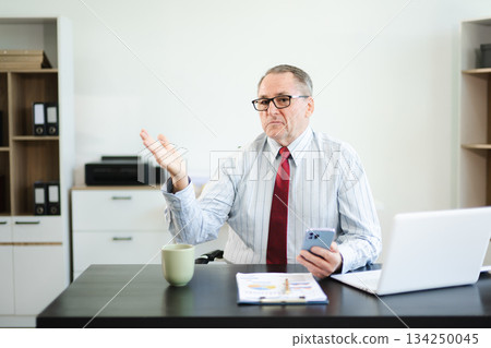 Frustrated senior businessman with smartphone gestures in confusion at office desk, reacting to bad news, tech issue, or business problem with laptop and reports. Frustrated senior businessman with smartphone gestures in confusion at office desk, reacting to bad news, tech issue, or business problem with laptop and reports. 134250045