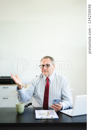 Frustrated senior businessman with smartphone gestures in confusion at office desk, reacting to bad news, tech issue, or business problem with laptop and reports. Frustrated senior businessman with smartphone gestures in confusion at office desk, reacting to bad news, tech issue, or business problem with laptop and reports. 134250046