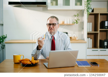 Mature Businessman using laptop while sitting at home. man sitting in kitchen Mature Businessman using laptop while sitting at home. man sitting in kitchen 134250047